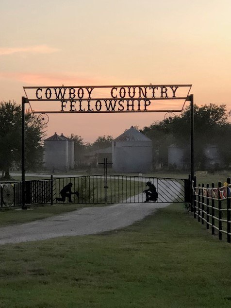 Entrance gate to Cowboy Country Fellowship with COWBOY COUNTRY FELLOWSHIP sign and kneeling cowboy silhouettes at dusk