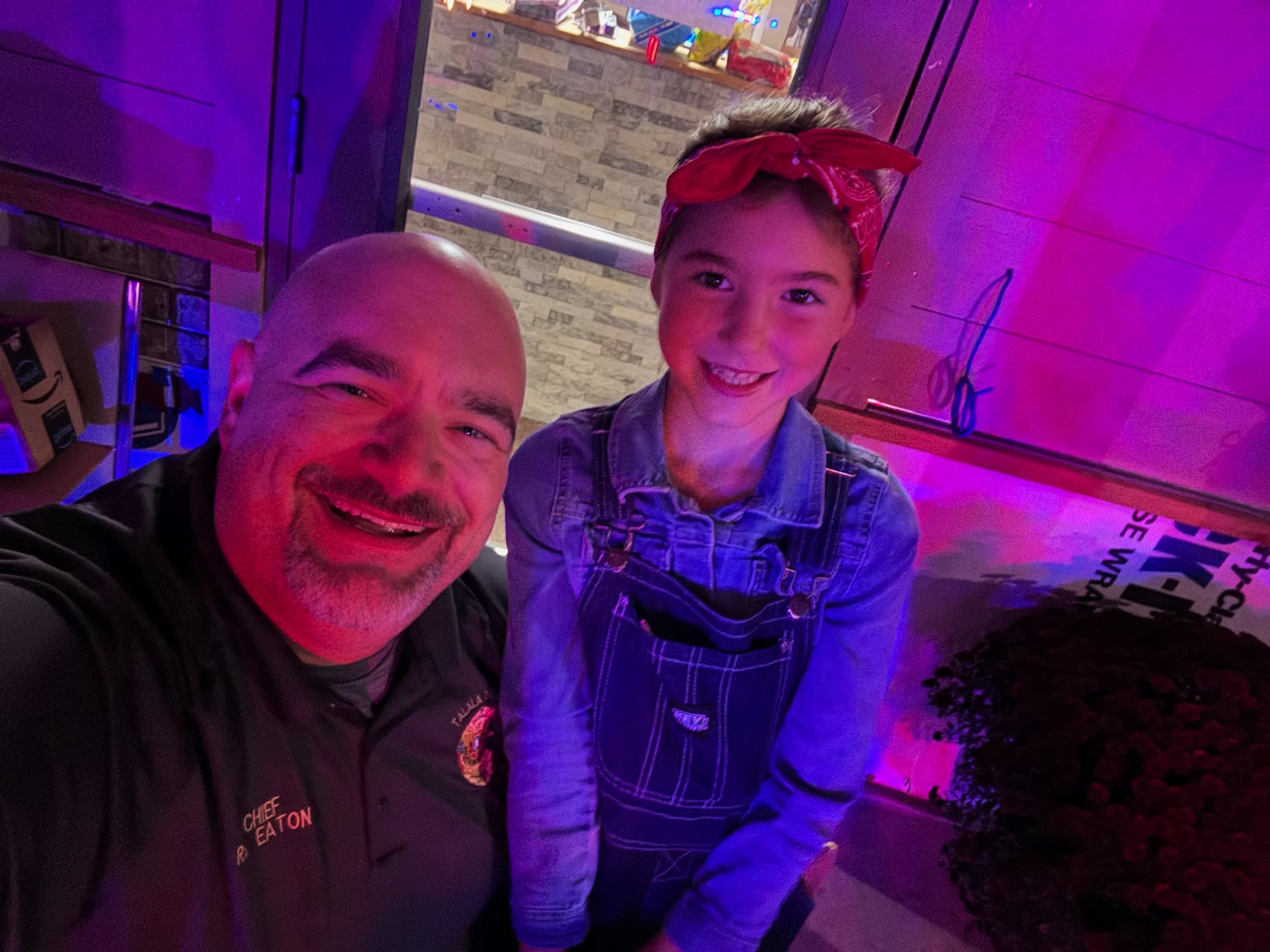 Chief R. Eaton in a Talala Police polo selfie with a smiling young girl in denim overalls and a red bandana under purple event lighting