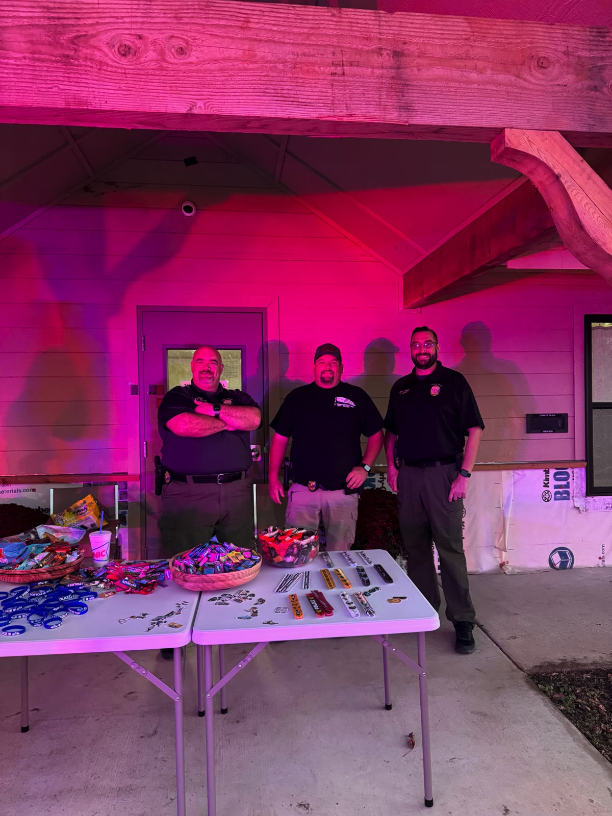 Chief R. Eaton, Asst. Chief Travis Linzy, and an officer behind candy and prize tables under pink event lighting