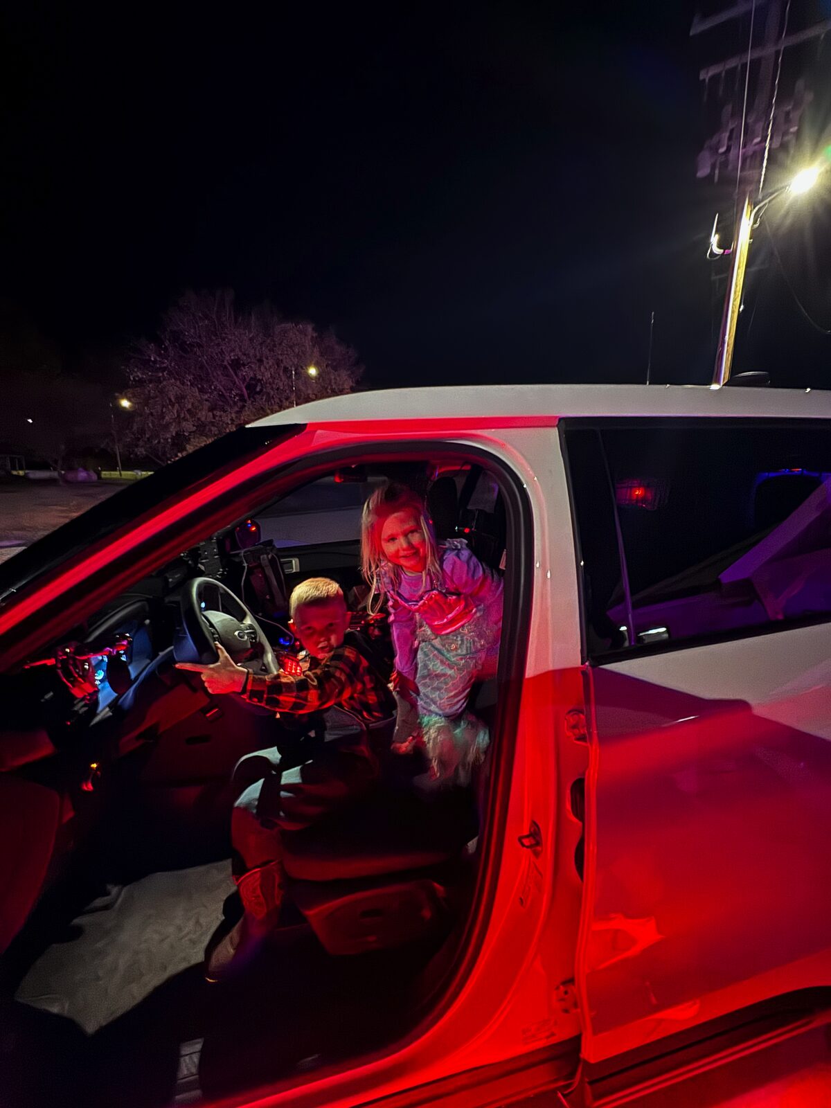 Two children exploring a Talala PD cruiser at night under pink emergency lighting, one in a mermaid costume