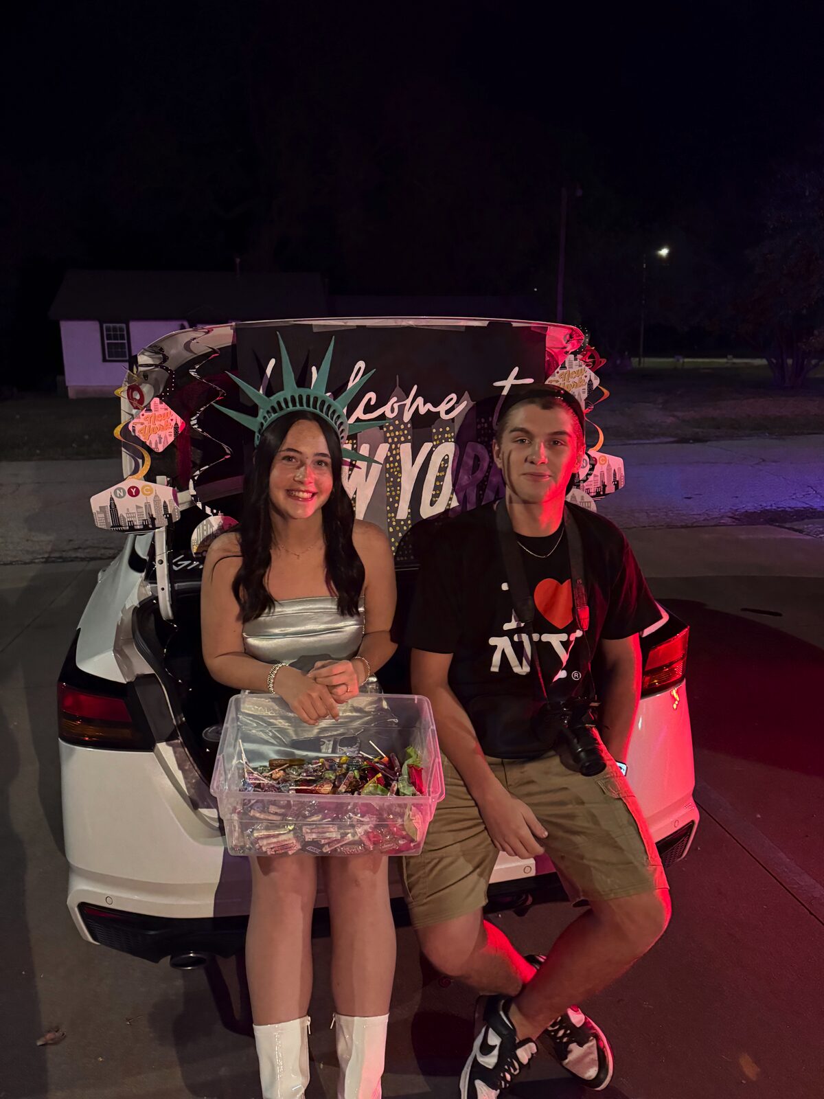 Young community members in an open car trunk decorated Welcome to New York for trunk-or-treat, one in a Statue of Liberty crown holding candy